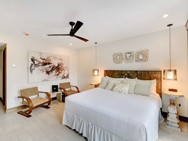 This image shows a modern bedroom with a large bed, two chairs, a ceiling fan, and wall art. The decor is minimal and elegant.