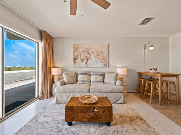 A cozy living room with a large window, a beige sofa, a wooden coffee table, a painting, lamps, and a small bar area with stools, all well-lit.