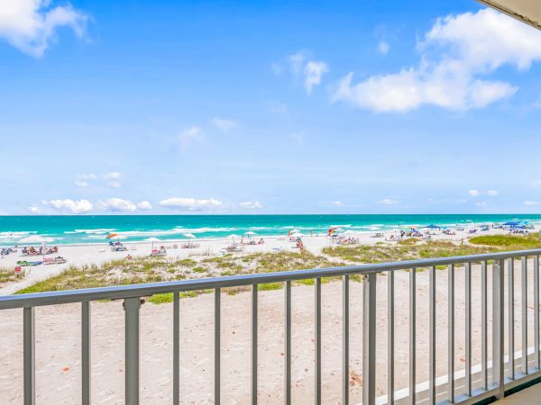 A balcony view of a sandy beach with people, umbrellas, and the ocean under a bright blue sky with some clouds, ending the sentence.