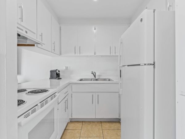 A modern white kitchen with cabinets, a sink, stove, refrigerator, and a small coffee machine on the counter. The floor has beige tiles.