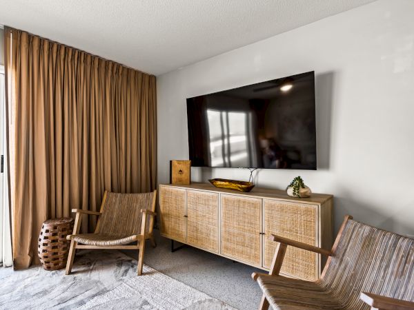 The image shows a modern living room with a wall-mounted TV, two wicker chairs, a light brown rug, and a wooden cabinet under the TV.