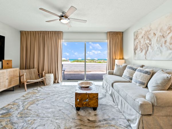 A modern living room with a large sofa, patterned rug, wooden furniture, and sliding doors opening to a beach view with blue skies.
