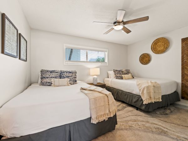 A cozy bedroom with two beds, stylish decor, a ceiling fan, wall art, and a large mirror. A window provides natural light.
