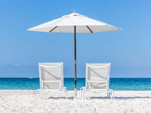 Two empty white beach chairs under a large umbrella on a sunny sandy beach, calm blue ocean in the background.