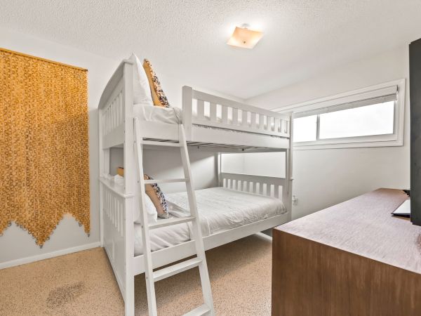 The image shows a small bedroom with a white bunk bed, a dresser with a TV, a window, and a wall hanging made of small yellowish items.