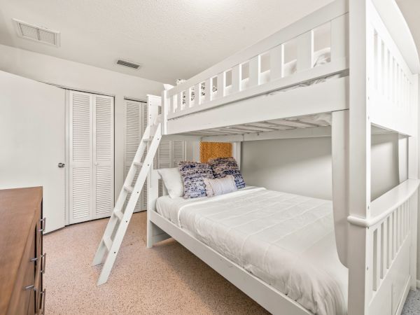 A bedroom with white bunk beds, featuring a ladder and blue-patterned pillows, beside a wooden dresser and partially open closet doors.