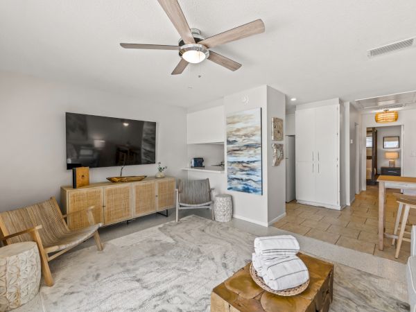 A modern living room with a ceiling fan, wall-mounted TV, wooden furniture, and coastal decor. The image includes a hallway and a small dining area.