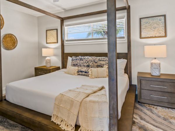 A cozy bedroom with a wooden four-poster bed, two bedside tables with lamps, and decorative wall art. The room has a serene, neutral palette.