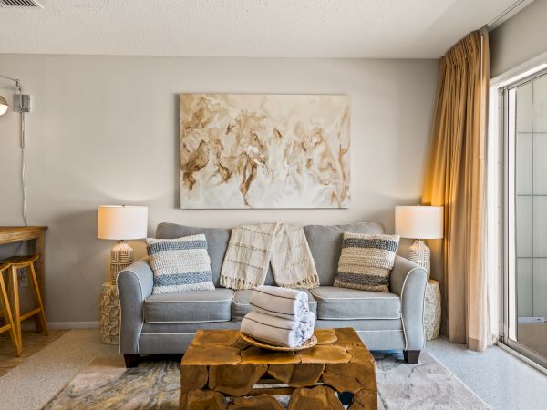 A cozy living room with a gray sofa, abstract art, two lamps, a unique wooden coffee table, rolled towels, and a small bar area to the left, ending the sentence.