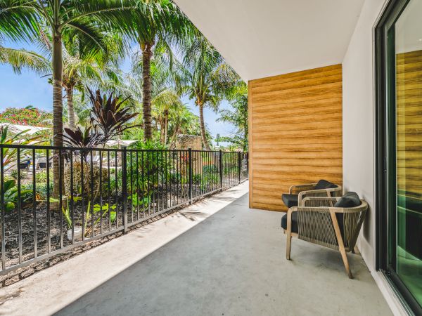 A tropical patio with two chairs, wooden wall accents, potted plants, and a view of lush greenery, enclosed by a black railing.