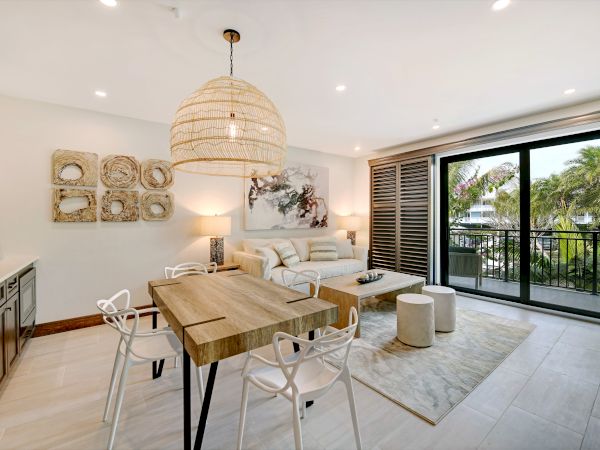 A modern open-concept living area with dining table, sofa, artwork, pendant lamp, and large sliding doors leading to a balcony with greenery outside.