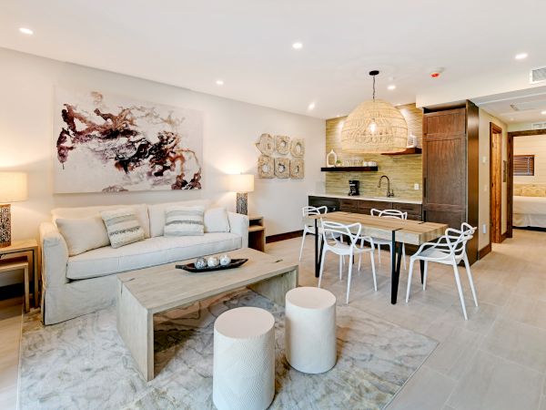 The image shows a modern living room and kitchen area with stylish furniture, a couch, dining table, and wall art, leading to a bedroom.