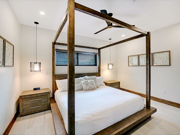 A modern bedroom with a wooden canopy bed, white bedding, and nightstands. It features hanging lights, framed art, and a ceiling fan on the light wood floor.