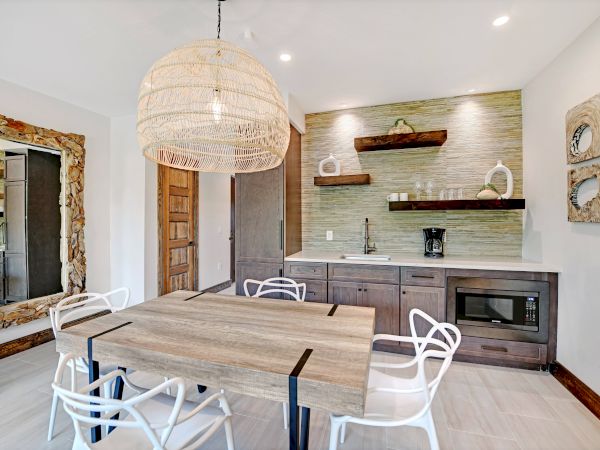 A modern dining room with a wooden table, white chairs, a large woven pendant light, wall art, and a kitchenette with shelves and a microwave.