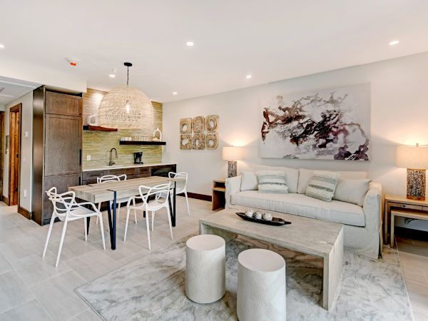 A modern living area with a sofa, dining table, and kitchenette. Decor includes abstract art, pendant light, and stools on a rug, ending the sentence.