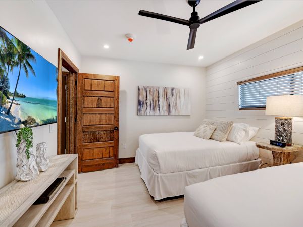 A modern bedroom with two beds, a wall-mounted TV displaying a beach scene, a ceiling fan, and stylish decor, ending the sentence.