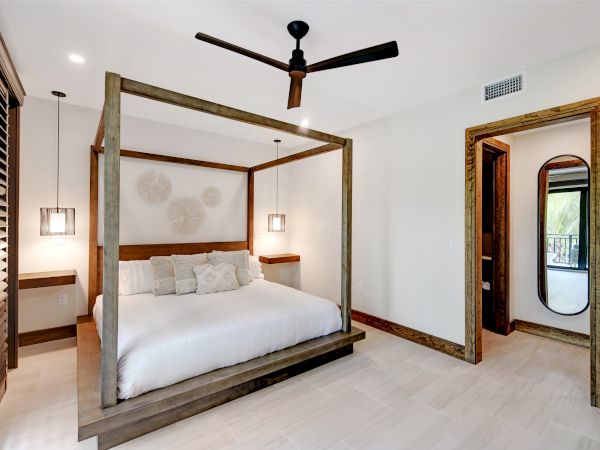 A minimalist bedroom with a canopy bed, modern ceiling fan, wall art, bedside tables with lamps, and a doorway leading to another room.