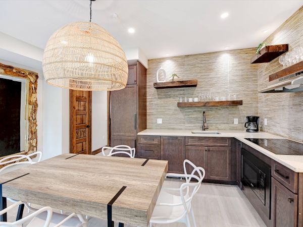 Modern kitchen with wooden cabinets, large pendant light, dining table, white chairs, shelves, and built-in appliances, featuring a rustic door.