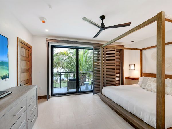 A modern bedroom with a canopy bed, ceiling fan, flat-screen TV, dresser, and sliding doors leading to a balcony with a tropical view.