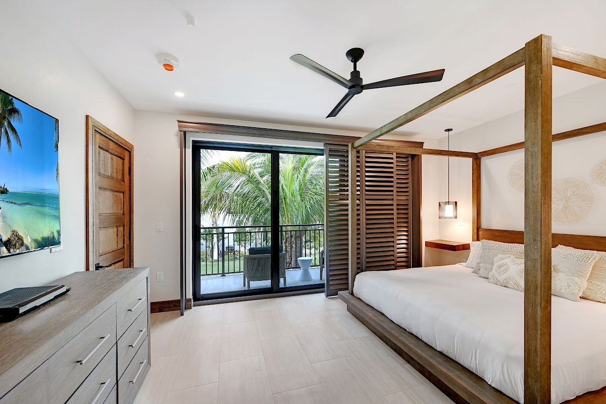 A bedroom with a wooden four-poster bed, ceiling fan, dresser with a TV, and glass doors leading to a balcony with a tropical view.