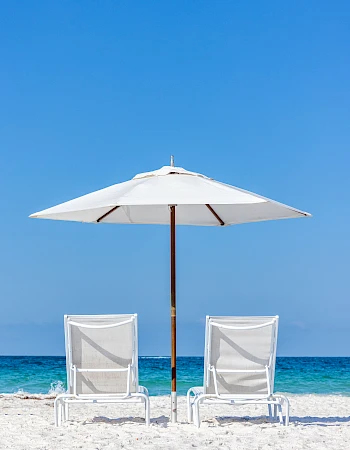 Two white beach chairs and an umbrella on a sandy beach with a blue sky and ocean in the background.