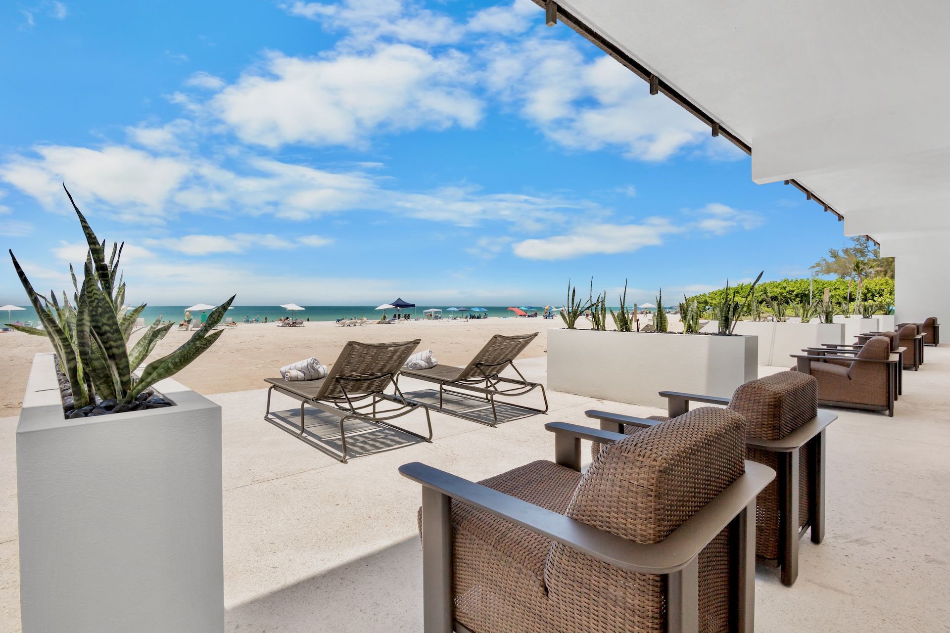 A sunny beachfront resort patio with wicker lounge chairs, planters, and umbrellas along a sandy beach under blue skies.