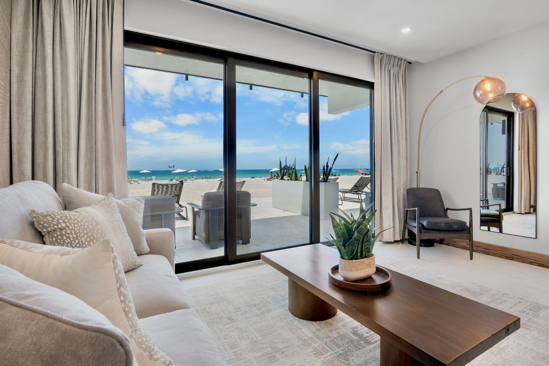 A bright, modern living room with beige sofa, wooden coffee table, and a large sliding glass door opening to a sunny beach view outside.