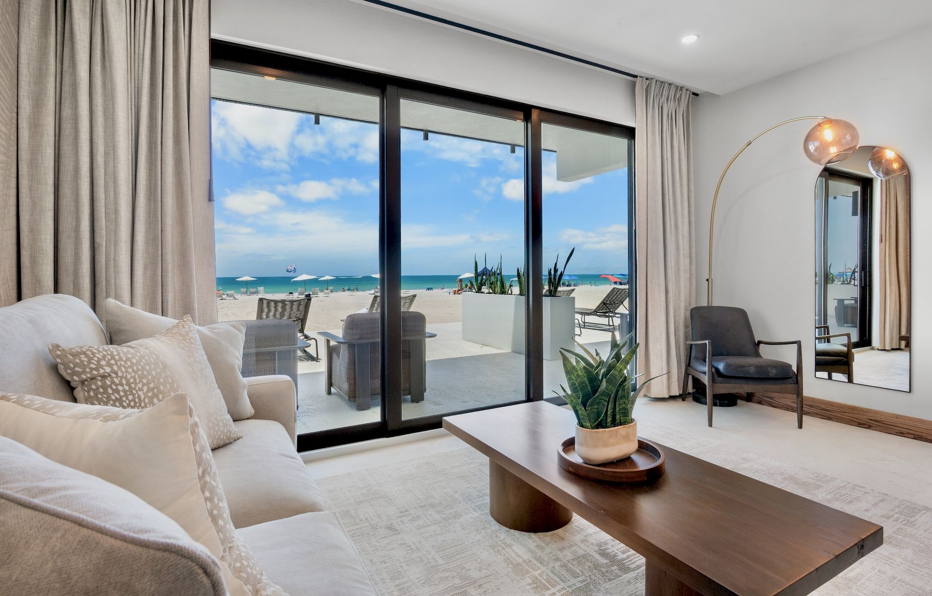 A bright, modern living room with beige sofa, wooden coffee table, and a large sliding glass door opening to a sunny beach view outside.