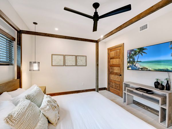 Modern bedroom with white bedding, wooden beams, ceiling fan, pendant light, and a TV showing a tropical beach scene.