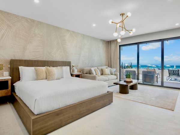 A modern hotel room with a large bed, a seating area, stylish decor, and floor-to-ceiling windows overlooking a beach and ocean, bright and airy.