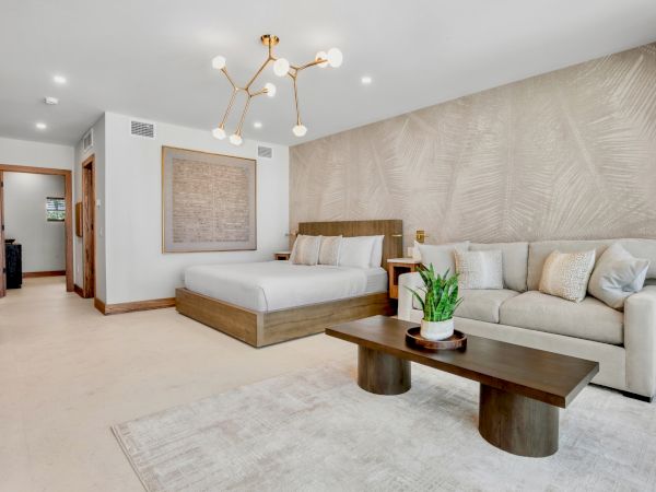 A modern hotel suite featuring a king bed, a cozy beige sofa, a wooden coffee table, neutral d&eacute;cor, wall art, and warm ambient lighting.