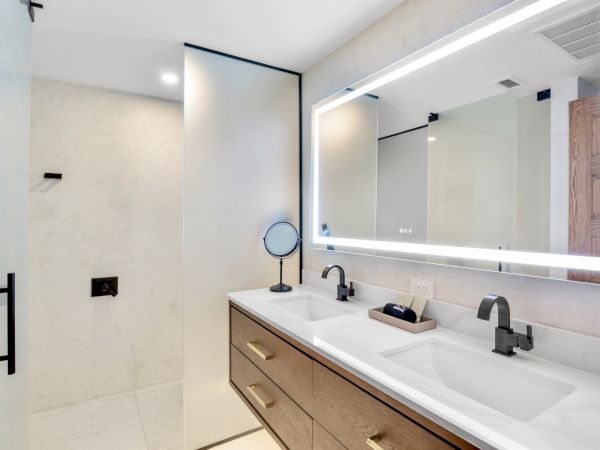 A modern bathroom with a double sink vanity, large mirror with backlit frame, light wood cabinets, and a glass-enclosed shower; sleek fixtures throughout.