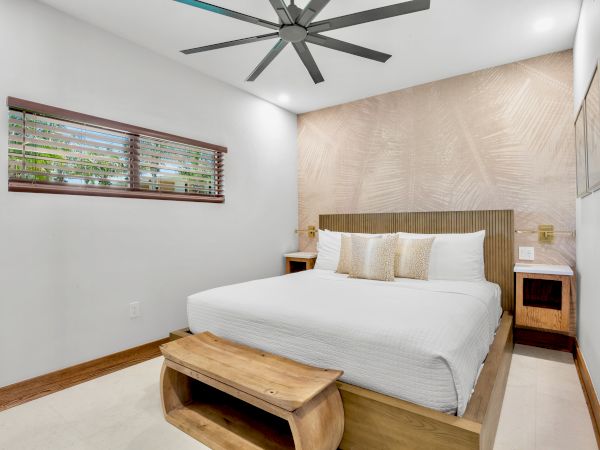 A modern hotel-style bedroom with a king bed, wooden headboard, two nightstands, a ceiling fan, a small bench at foot, and a window with blinds.
