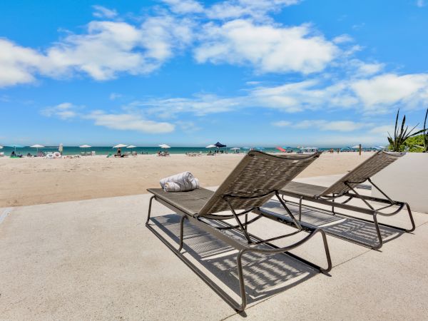 Two empty beach chairs face a sunny seaside with blue water, white clouds, and a wide sandy shore. End of sentence.