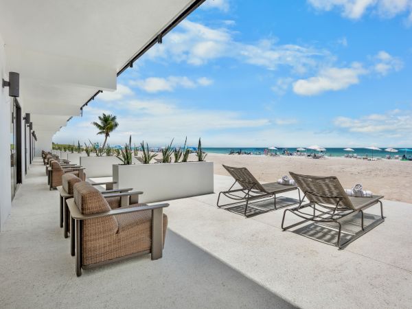 People lounge on water-view patio chairs and loungers outside a beachside building, with the ocean, palm trees, and blue sky in the background.
