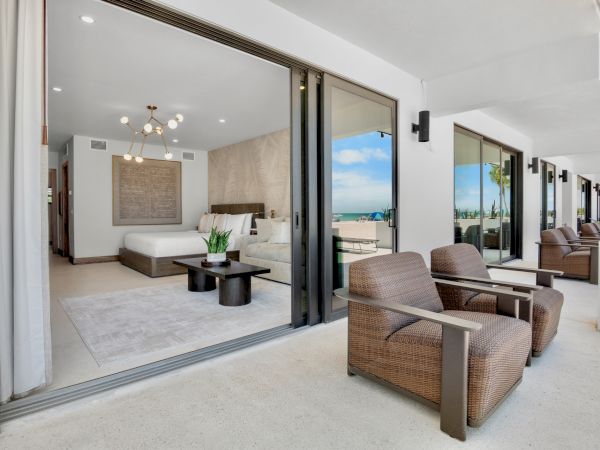 A modern hotel suite with a bright bedroom and a sliding glass door opening to a spacious balcony, featuring woven wicker chairs and ocean views.