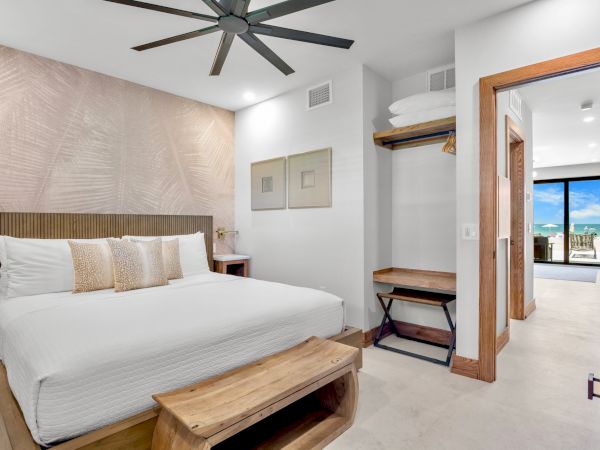 A modern hotel bedroom with a white king bed, beige upholstered headboard, matching pillows, wooden bench, ceiling fan, open closet, and a view toward a bright hallway and ocean-side balcony.