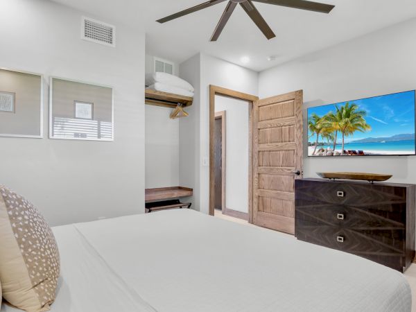A bright hotel-style bedroom with a bed, dresser, wall TV showing a beach scene, open closet, and wooden door in a clean, modern space.