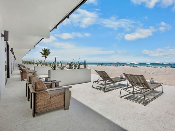 A beachside patio with wicker seating and loungers facing the sandy shore and blue ocean under a bright, partly cloudy sky.