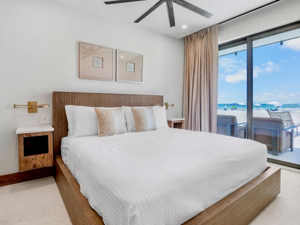 A bright, modern hotel bedroom with a large bed, neutral bedding, wooden furniture, and a balcony overlooking the ocean.