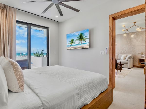 A bright, modern bedroom with a large bed, white bedding, sliding glass doors to a balcony, a wall-mounted tropical TV, and an open doorway to a living area.