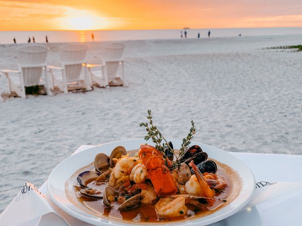 A plate of seafood pasta sits on a table by the beach at sunset, with the sea and sky in the background.