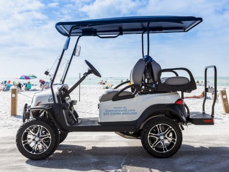 The image shows a golf cart parked on a paved area near a beach. Several people and umbrellas are visible on the sandy beach in the background.