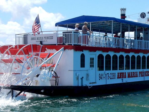 A red and white paddlewheel riverboat named "Anna Maria Princess" is cruising on the water with an American flag on its stern.