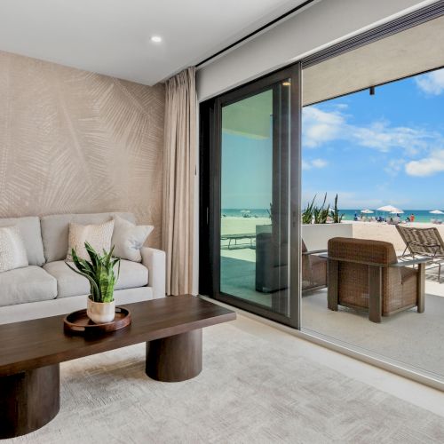 A stylish living room with a beige sofa, textured wallpaper, and a dark wooden coffee table, opens to a bright beach patio with lounge chairs.