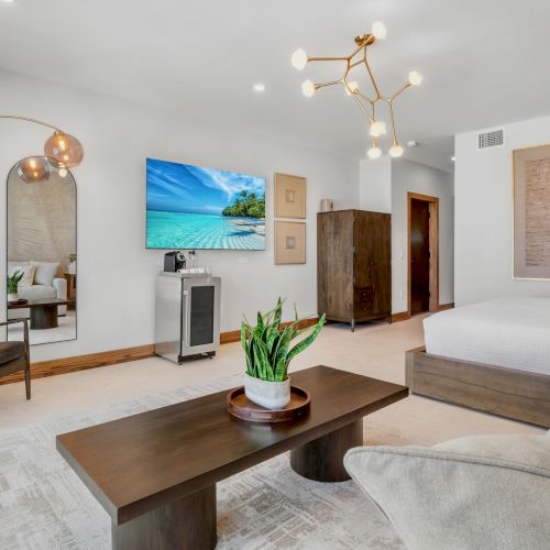 A cozy living room with a wood coffee table, beige sofa, armchairs, a TV mounted on the wall, and warm lighting.