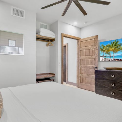 Bright modern bedroom with ceiling fan, bed, wood dresser/closet doors, and a wall TV showing a tropical beach scene.