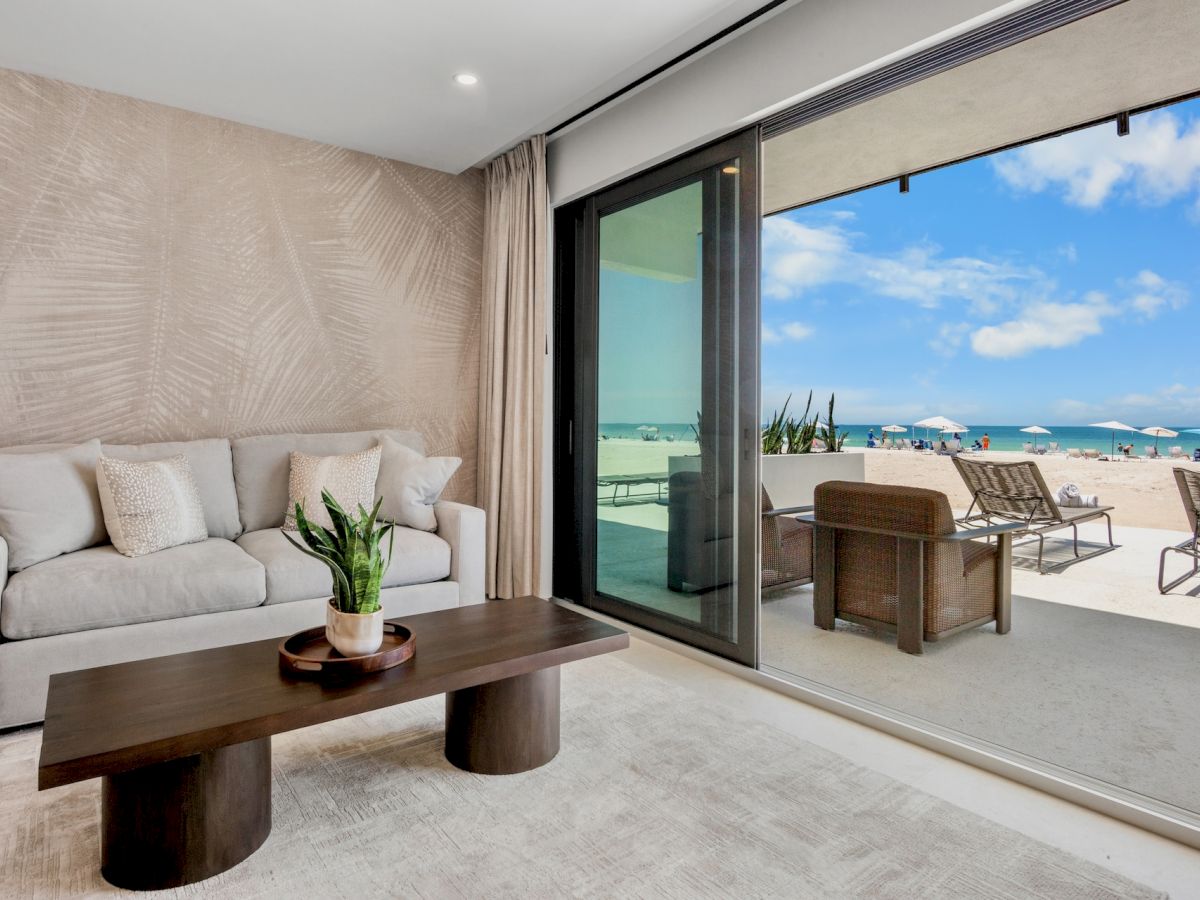 A modern living room with a beige sofa, textured wall, and a wooden coffee table, opening to a sunny beach terrace with lounge chairs.