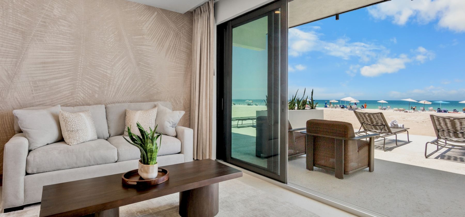 A modern living room with a beige sofa, textured wall, and a wooden coffee table, opening to a sunny beach terrace with lounge chairs.