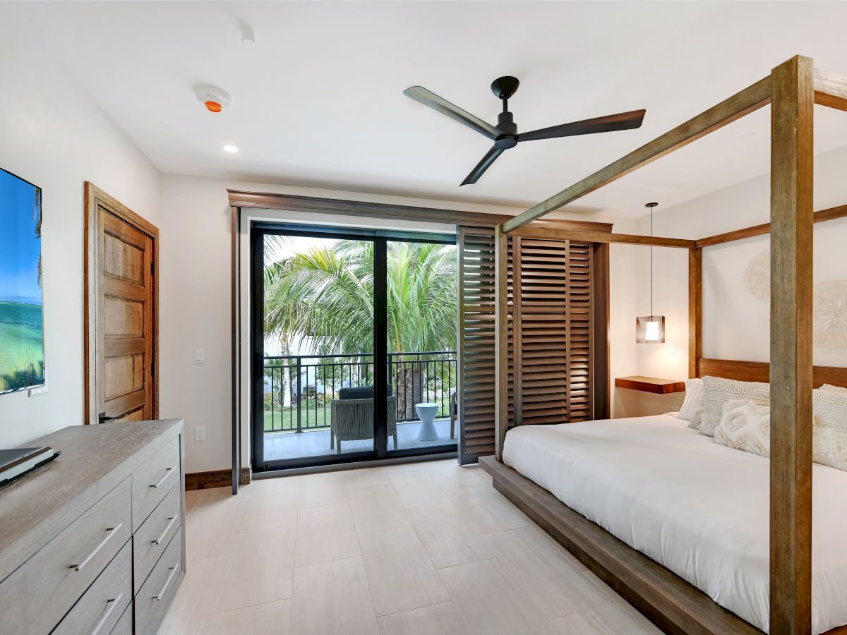 A bedroom with a wooden four-poster bed, ceiling fan, dresser with a TV, and glass doors leading to a balcony with a tropical view.